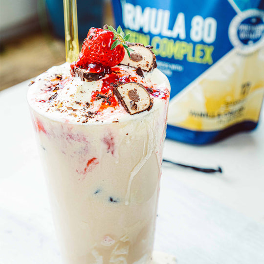 Ein Vanille Protein Shake mit Erdbeeren und Riegelsücken und im Hintergrund ein Proteinpulver Beutel